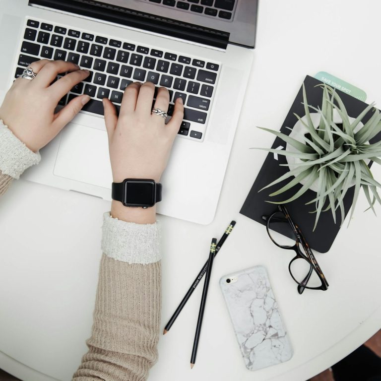 Buchhaltung auf Augenhöhe Hände tippen auf einer Tastatur, umgeben von Notizblock, Telefon und Pflanze.